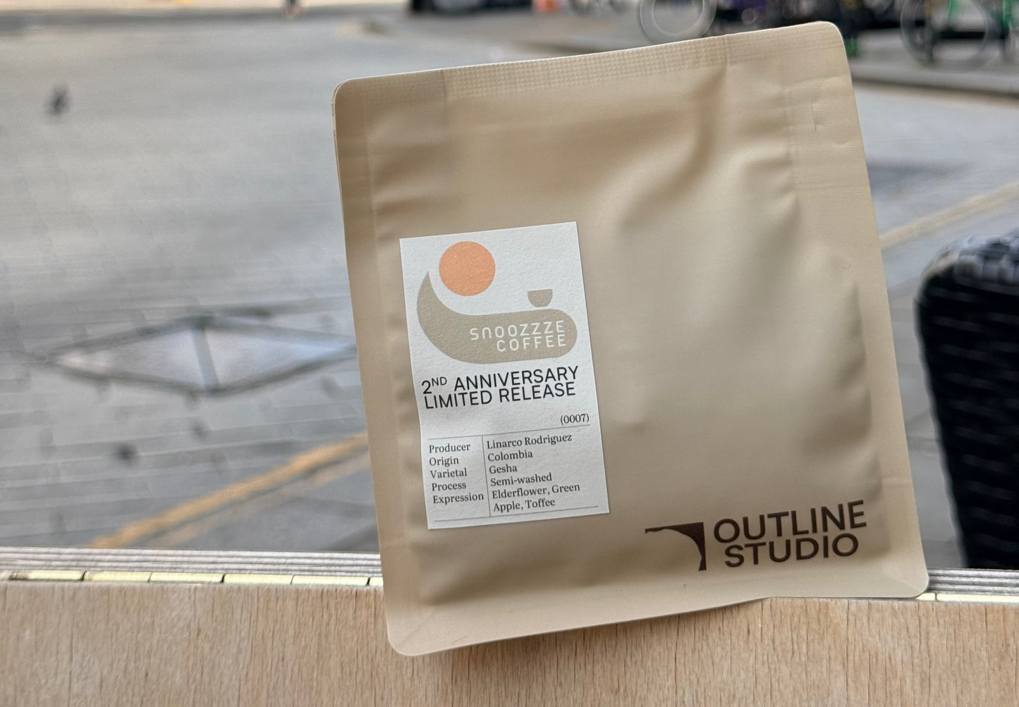 A stand-up pouch of Outline Studio coffee sits on a wooden ledge with a blurred city street scene in the background. The pouch is a warm beige/tan color with a white label that reads '2th Anniversary Limited Release' and shows the Outline Studio logo.