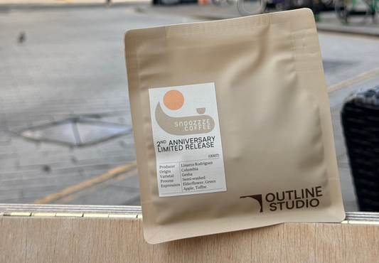 A stand-up pouch of Outline Studio coffee sits on a wooden ledge with a blurred city street scene in the background. The pouch is a warm beige/tan color with a white label that reads '2th Anniversary Limited Release' and shows the Outline Studio logo.